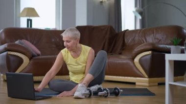 Orta yaşlı, olumlu gülümseyen bir sporcu kadın dizüstü bilgisayarla oturma odasındaki egzersiz paspasına oturmuş mesajlaşıyor. Kafkasyalı bir kadının evde mola eğitimi alırken geniş açılı bir portresi.