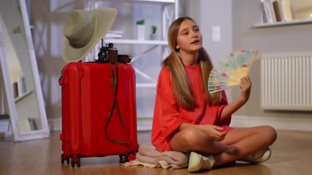 自信的少女坐在红色旅行袋旁 手拿着扇子等待 广博的照片描绘了一个充满自信的白人青少年正准备在室内旅行 旅游业与青少年 — 图库视频影像