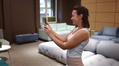 Dükkanda yeni mobilyaların fotoğrafını çeken gülümseyen kadın sosyal medya kuryesine fotoğraf yolluyor. Kendine güveni tam beyaz bir alıcı dükkanda yeni kanepe seçiyor ve tavsiye istiyor.