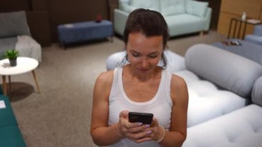 Özümsenmiş beyaz bir kadının akıllı telefondan dokunmatik ekranda yazıp başka yöne bakmasının yüksek açılı görüntüsü. Düşünceli bayan müşterinin portresi mobilya salonundaki yeni kanepeyi internetten seçiyor.