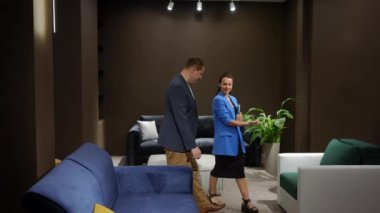 Geniş açılı profesyonel kadın dijital tabletle mobilya salonuna giriyor ve erkek işaret ediyor. Uzman Kafkasyalı satış müdürü, içerideki koltuğu seçen müşteriye yeni kanepe reklamı yapıyor.