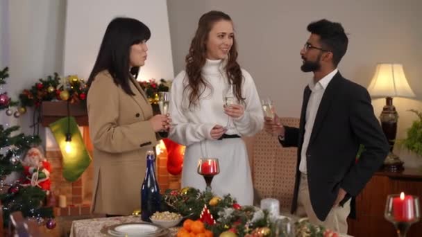 三个快乐的跨国朋友站在室内圣诞餐桌前祝酒 积极自信的中东男人亚洲和高加索女人叮咬香槟酒杯 微笑着说话 — 图库视频影像