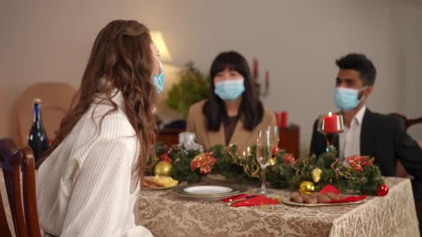 Covid 19顔マスクの悲しい白人女性は バックグラウンドでクリスマスディナーテーブルに座って話している友人とカメラの目撃を見回します コロナウイルスの新年にポーズをとって落ち込んでいる女性 — ストック動画