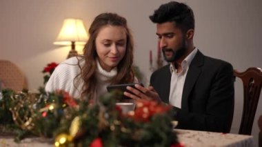 Yeni yıl arifesinde yemek masasında oturan konsantre beyaz kadın ve Orta Doğulu erkek akıllı telefondan sosyal medyayı kaydırıyor. Rahatlamış genç çift Noel 'i birlikte kutluyor.