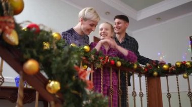 Heyecanlı erkek kadın ve genç kız Noel sabahı evde süslü tırabzanların önünde dikilip aşağıya bakıyor ve konuşuyorlar. Mutlu Kafkasyalı baba, anne ve kız mutlu bir yeni yıl geçirdiler.