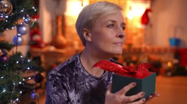 Yakın plan, çekici yetişkin bir kadın Noel gecesi hediye kutusuyla rahat bir oturma odasında oturuyor. Gülümseyen güzel beyaz kadın evde yeni yıl arifesinde mucize diliyor.