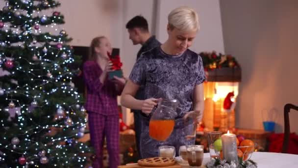 快乐的母亲把果汁倒在玻璃杯里 父亲和女儿站在圣诞树旁笑着 积极的白人妇女在家里和家人一起准备新年大餐 — 图库视频影像