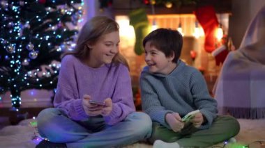 Neşeli çocuk ve kız, yeni yıl arifesinde akıllı telefonlarda dedikodu yapan sosyal medyaya gülüyorlar. Neşeli beyaz kardeşlerin geniş açılı portresi Noel gecesi evde eğleniyorlar.