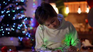 Oturma odasındaki Noel ışıklarının arka planında oturan konsantre çocuk portresi. Kafkasyalı rahat bir genç Noel Baba 'dan yeni yıl arifesinde mucize istiyor.