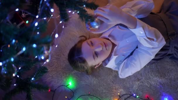 尽收眼底的少女躺在圣诞树下挂着装饰品微笑 在室内享受节日准备的快乐 轻松的白人青少年的高角肖像 — 图库视频影像