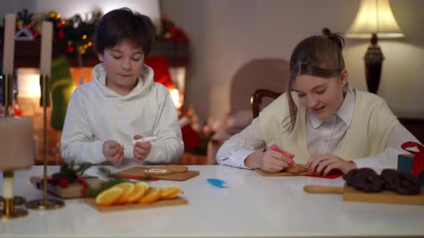 充满自信的男孩和女孩坐在客厅的桌子旁装饰圣诞饼干 快乐的白人兄弟姐妹为新年做美味的传统甜食的肖像 — 图库视频影像