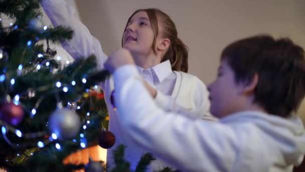 スローモーションの笑顔で自宅でクリスマスツリーを飾る積極的な男の子と女の子 新年の準備を楽しむ幸せな白人の妹と弟屋内で前夜 — ストック動画