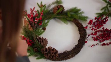 Yüksek açılı el yapımı Noel çelengi ve kırmızı karlı böğürtlen takan bir kadın eli. Masada yeni yıl süsü veren zayıf, genç, beyaz bir kadının omzundan ateş etmek.