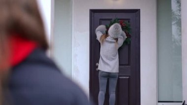 Arkadan bakan genç bayan, Noel çelengi ile kapıyı dekore ederek komşusunu selamlıyor. Neşeli Kafkasyalı güzel kadınlar yeni yıl arifesinde dışarıda buluşmayı kucaklıyor.