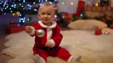 Noel topuyla oynayan özümsenmiş bebek Noel Baba kırmızı çantayla oturma odasına giriyor. Tasasız beyaz çocuğun portresi. Yeni yıl arifesinde evdeki sevimli çocuk.