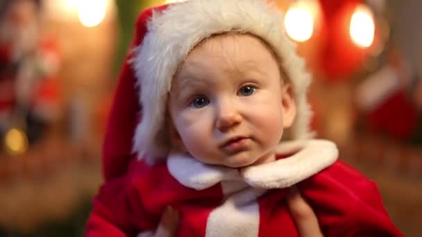 サンタクロースの衣装に身を包んだ明るい赤ちゃんが クリスマスライトを背景に笑顔でカメラを見ている様子を間近で見ることができます 幸せな正白人の幼児Posingで家に上の新年Eve — ストック動画
