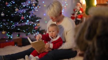 Noel Baba kostümlü hiperaktif bir kız bebek bebek için kitap okuyan bir kadın olarak el kol hareketi yapıyor. Kafkasyalı kaygısız çocuk pozitif annesiyle evde dinleniyor.