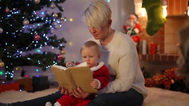 新年の若い美しい女性との本を読んでキュリオかなり赤ちゃんの女の子は自宅で前夜 クリスマスに叔母と屋内で趣味を楽しむかわいい白人の幼児の肖像画 知性と幼児期 — ストック動画