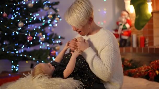 クリスマスの前夜にリビングルームでかわいい娘の小さな足にキスをする興奮した愛する母親 ポジティブな白人の美しい女性の肖像とともにかなり幼児で新年屋内 — ストック動画