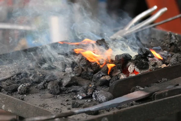 Coal, smoke and fire on foreground and tools for a blacksmith on background, close-up