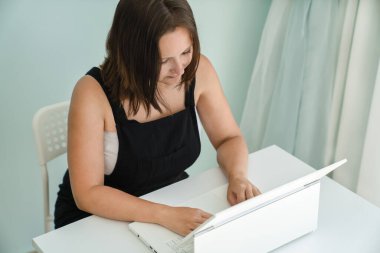 Genç bir kadın evindeki masasında dizüstü bilgisayarının önünde oturuyor. Serbest meslek ve uzaklık eğitimi kavramı, yan görüş