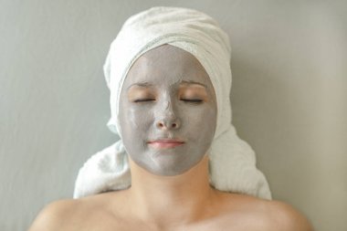 Yüzünde kil maske ve kafasında havlu olan genç bir kadın türban şeklinde gözleri kapalı yatıyor ve yüz bakımı için kozmetik spa tedavisinden hoşlanıyor. Yüz yakın çekim, üst görünüm.