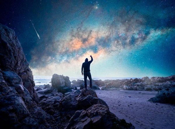man standing on the rock, back view,  with raised hand staring the Milky Way and stars over the ocean