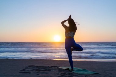 Bilinmeyen Latin kadın gün batımında plajda geriniyor ya da yoga yapıyor.