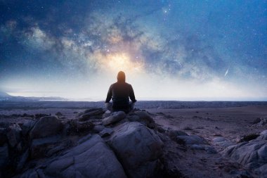 Bir insanın silueti dağda oturmuş Samanyolu 'nun altında meditasyon yapıyor ve yıldızlı bir gecede geriye bakıyor.
