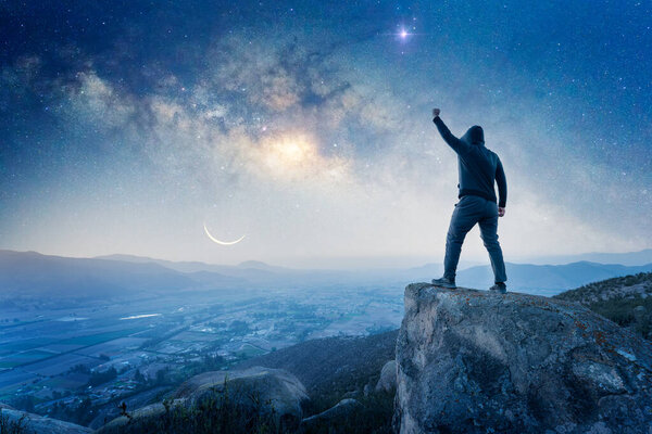 man standing on the top of the mountain, back view, with a hand up celebrating and Milky Way over the valley	