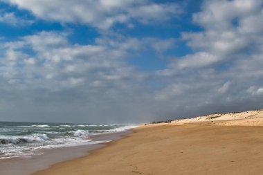 Bulutlu bir gökyüzünün altında güneşli kumulları olan ıssız bir sahil.