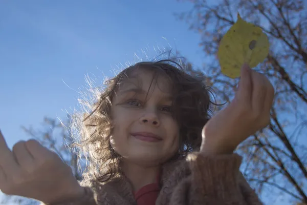 Uma menina sorridente contra o céu azul segura uma folha de outono em suas mãos. — Fotografia de Stock