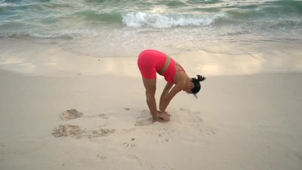 Mujer joven en traje deportivo practicando yoga en la playa al amanecer cerca del mar — Vídeos de Stock