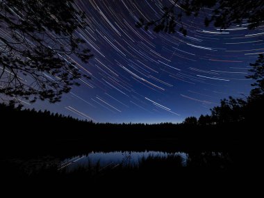 Orman göletinin yukarısındaki yıldız izleri. Bir uçak gece gökyüzünde kendi yolunu çizer..