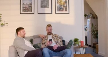 Bir LGBTQI 'nin ön görüntüsü. Oturma odasında oturan aynı cinsiyetten erkek çift. Bir adam ortağına akıllı telefonunu gösteriyor..