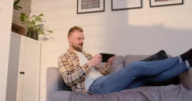 Oturma odasında kanepede uzanmış, dinlenen genç bir adamın yan görüntüsü. Akıllı telefonunu online bir oyun oynamak için kullanıyor..