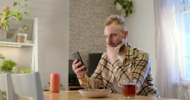 Oturma odasındaki masada oturan genç bir adamın yan görüntüsü, rahatlarken, akıllı telefonunu kullanıyor ve yanında bir tabak makarna var..