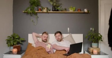 Bir LGBTQI 'nin ön görüntüsü. Aynı cinsiyetten erkek çift yataklarında dinleniyor. Laptop ve akıllı telefon kullanıyorlar..