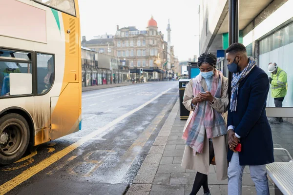 Pareja Esperando Que Autobús Weraing Revestimientos Faciales Mirando Aplicación Móvil —  Fotos de Stock