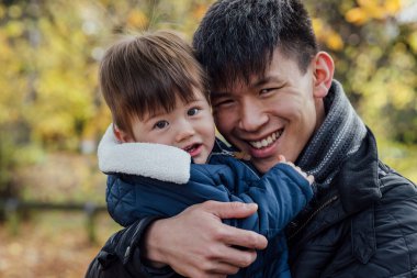 A young man carrying his baby boy in his arms while standing outside and embracing him in nature in Northumberland, North East England. They are both looking at the camera and smiling.