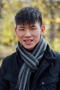 A close-up portrait of a young, Chinese man standing outdoors in nature in Northumberland, North East England during Autumn. He is smiling while looking at the camera.