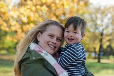 A mid adult woman carrying her baby boy in her arms while standing outside and embracing him in nature in Northumberland, North East England. They are both looking at the camera and smiling.