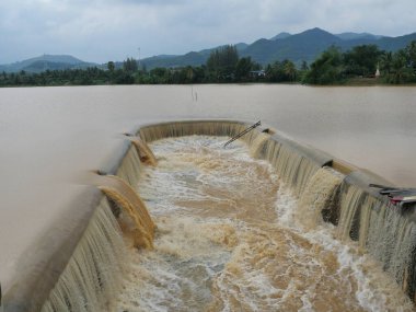 Barajda taşan kahverengi su, arka planda dağ ve yeşil orman, yağmur mevsiminde sel, Tayland