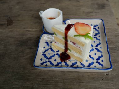 Çilekli ve kivili meyve dilimli kırmızı renkli şurup ve kremalı kek gri ahşap masadaki seramik tepside, beyaz fincanda sıcak espresso kahvesi