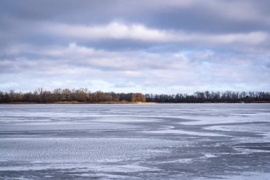 Karla kaplı donmuş bir nehir, donmuş bir nehir kıyısı, nehrin diğer tarafındaki bir adaya güneş ışığı düşüyor.