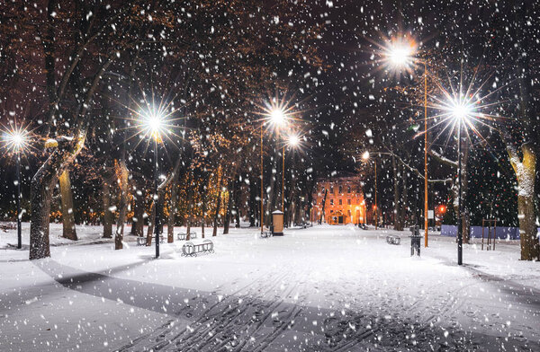 Snowfall in a winter park at night with christmas decorations, glowing lanterns, pavement covered with snow and trees.