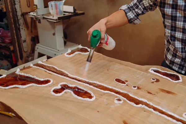 Carpenter removing air bubbles with blowtorch from epoxy table. Process of making a craft resin and wood table