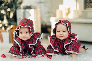 Cute identical twin babies in beautiful red dresses smiling to the camera and lying on the floor and playing near Christmas tree and gifts clipart