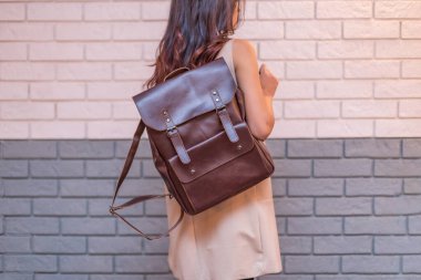 Woman holding brown leather backpack in the hand on the brick wall background. Unisex bag clipart