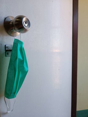 green surgical face mask hanging on a silver doorknob of a white wooden door, symbolizing health safety, hygiene, and daily pandemic precautions in a home or medical setting